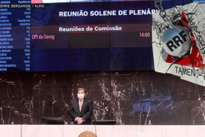 Imagem Acessível: Reunião de Sessão Legislativa no plenário da ALMG, na mesa há um homem branco em pé, que preside a sessão, o deputado Agostinho Patrus. Atrás desta mesa está o painel de votações. Em primeiro plano, no topo, há uma intervenção gráfica com a representação de um muro com o mapa de Minas Gerais sendo destruído por uma bola de demolição, que está com o rótulo de RRF (Regime de Recuperação Fiscal)