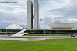 Foto do Congresso Nacional, local que sedia as duas casas do Poder Legislativo Brasileiro – no centro há duas torres, ao lado de cada uma delas há duas cúpulas, uma côncava (Senado) e outra convexa (Câmara). O céu está nublado e, ao redor da edificação, há uma grande área com gramado verde.