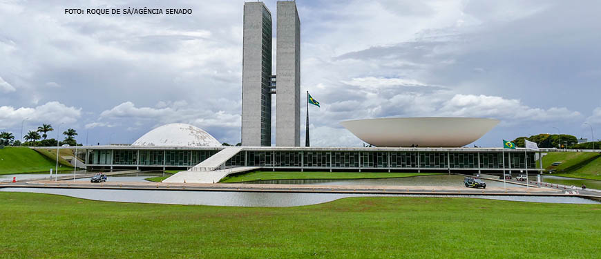 Foto do Congresso Nacional, local que sedia as duas casas do Poder Legislativo Brasileiro – no centro há duas torres, ao lado de cada uma delas há duas cúpulas, uma côncava (Senado) e outra convexa (Câmara). O céu está nublado e, ao redor da edificação, há uma grande área com gramado verde.