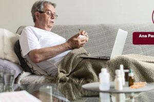 Fotografia mostra um idoso sentado em um sofá, de óculos e camisa branca, com um laptop no colo. Ele parece estar realizando alguma atividade no laptop.