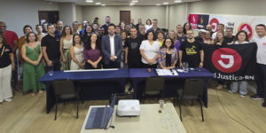 A imagem mostra uma grande reunião de representantes do SINJUS e da FENAJUD em uma sala de conferência iluminada. Aproximadamente 40 pessoas estão reunidas, em pé e sorrindo para a foto, atrás de uma longa mesa com toalha azul escura. Faixas e banners dos sindicatos estão fixados na parede ao fundo.