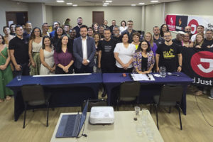 A imagem mostra uma grande reunião de representantes do SINJUS e da FENAJUD em uma sala de conferência iluminada. Aproximadamente 40 pessoas estão reunidas, em pé e sorrindo para a foto, atrás de uma longa mesa com toalha azul escura. Faixas e banners dos sindicatos estão fixados na parede ao fundo.