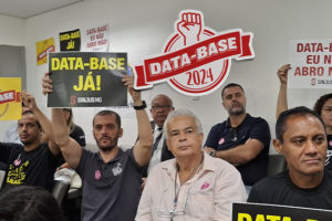 A imagem mostra servidores e sindicalistas em um ambiente fechado, segurando cartazes com frases como “DATA-BASE JÁ!” e “DATA-BASE EU NÃO ABRO MÃO!”. Alguns estão sentados, outros de pé, todos com expressões sérias e determinadas, demonstrando mobilização pela reposição salarial de 2024. Ao fundo, há cartazes e banners fixados nas paredes, reforçando a pauta do movimento.