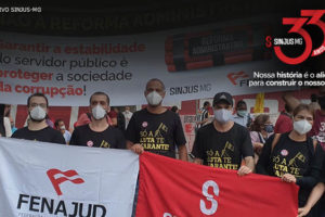 Grupo de homens e mulheres (dirigentes do SINJUS e servidores) protestam com faixas contra a Reforma Administrativa. Acima da foto, logo comemorativa, composta pelo número 33 em vermelho e do seguinte texto: Sinjus-MG 33 anos - Nossa história é o alicerce para construir o nosso futuro.