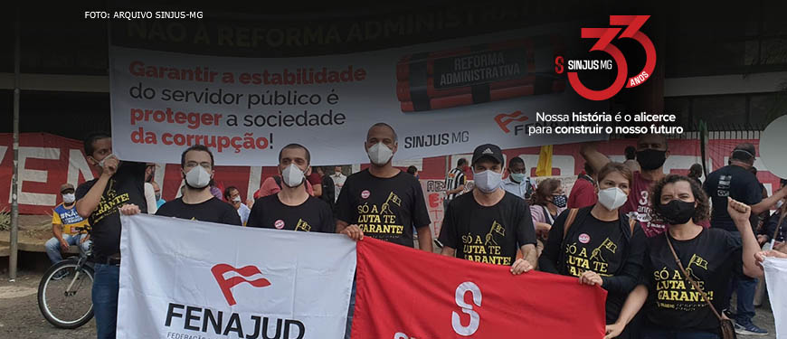 Grupo de homens e mulheres (dirigentes do SINJUS e servidores) protestam com faixas contra a Reforma Administrativa. Acima da foto, logo comemorativa, composta pelo número 33 em vermelho e do seguinte texto: Sinjus-MG 33 anos - Nossa história é o alicerce para construir o nosso futuro.