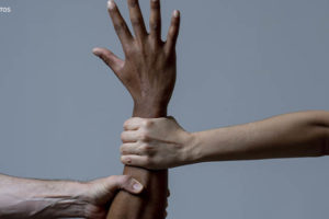Imagem Acessível: Um braço negro levantado com a mão aberta para o alto é puxado por dois braços de pessoas brancas, como se impedissem o movimento da pessoa negra.