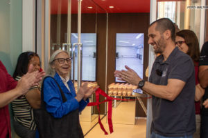 A imagem mostra um grupo de pessoas aplaudindo e comemorando, na cerimônia de corte de fita, indicando a inauguração do Espaço Lea Leda. A filiada Nolda Armani, uma senhora de cabelos brancos, aparece ao lado do coordenador-geral, Alexandre Pires, um homem branco, de cabelos pretos. Ele veste camisa azul escura e calça jeans.