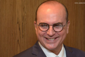 Foto de um homem branco, calvo e de olhos azuis, ele está sorridente e veste um terno azul marinho.