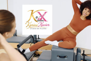 Imagem Acessível: Card de divulgação de convênio do SINJUS-MG com o Karine Xavier Studio de Pilates. A imagem mostra um estúdio de pilates com equipamentos . Em primeiro plano, desfocada à esquerda, aparece parte de uma pessoa sentada em um equipamento, observando a aula. Ao centro, está o logotipo do Karine Xavier Studio de Pilates. À direita, uma mulher negra, de cabelos cacheados volumosos, sorri enquanto realiza um exercício de alongamento lateral sobre um equipamento de pilates. Ela veste conjunto esportivo terracota de manga longa e calça legging, com meias brancas. Conteúdo textual: PILATES - Cuide do corpo e da mente com o novo convênio do SINJUS.