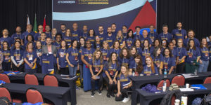 rupo de aproximadamente 60 pessoas, homens e mulheres, posando para foto coletiva em frente a um painel do 13º Consinjus – Congresso dos Servidores da Justiça de Minas Gerais. Todos vestem camisetas azuis do evento, com frases em amarelo, e alguns usam crachás de identificação. Ao fundo, há bandeiras do Brasil, de Minas Gerais e outra bandeira estadual. As pessoas estão sorridentes e distribuídas em duas fileiras, em um auditório com mesas e cadeiras vermelhas e pretas.