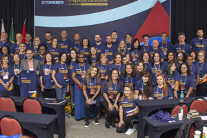 rupo de aproximadamente 60 pessoas, homens e mulheres, posando para foto coletiva em frente a um painel do 13º Consinjus – Congresso dos Servidores da Justiça de Minas Gerais. Todos vestem camisetas azuis do evento, com frases em amarelo, e alguns usam crachás de identificação. Ao fundo, há bandeiras do Brasil, de Minas Gerais e outra bandeira estadual. As pessoas estão sorridentes e distribuídas em duas fileiras, em um auditório com mesas e cadeiras vermelhas e pretas.