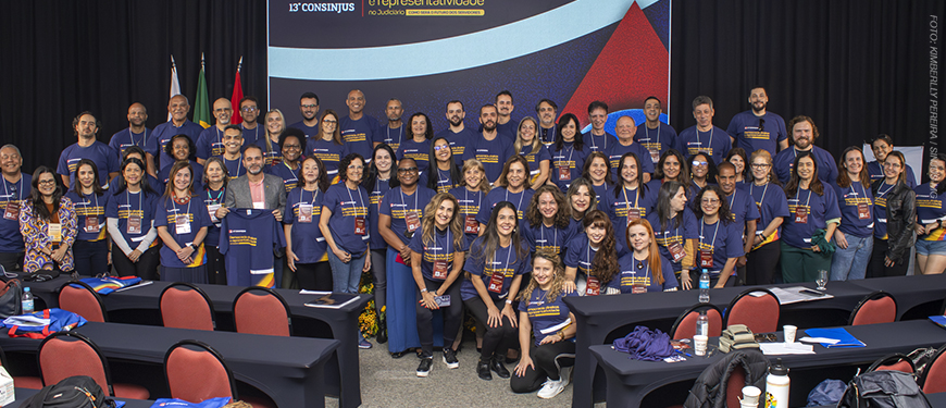 rupo de aproximadamente 60 pessoas, homens e mulheres, posando para foto coletiva em frente a um painel do 13º Consinjus – Congresso dos Servidores da Justiça de Minas Gerais. Todos vestem camisetas azuis do evento, com frases em amarelo, e alguns usam crachás de identificação. Ao fundo, há bandeiras do Brasil, de Minas Gerais e outra bandeira estadual. As pessoas estão sorridentes e distribuídas em duas fileiras, em um auditório com mesas e cadeiras vermelhas e pretas.