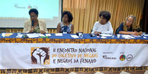 Imagem Acessível: A imagem mostra uma mesa de debates com quatro mulheres negras participando do II Encontro Nacional do Coletivo de Negras e Negros da Fenajud. Elas estão sentadas atrás de uma mesa coberta com tecido estampado e uma faixa que identifica o evento. Ao fundo, um telão projeta a identidade visual da atividade. O card traz o título: “SINJUS-MG reforça compromisso Antirracista em encontro nacional da FENAJUD”, destacando o compromisso da entidade com a pauta da igualdade racial.