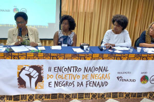 Imagem Acessível: A imagem mostra uma mesa de debates com quatro mulheres negras participando do II Encontro Nacional do Coletivo de Negras e Negros da Fenajud. Elas estão sentadas atrás de uma mesa coberta com tecido estampado e uma faixa que identifica o evento. Ao fundo, um telão projeta a identidade visual da atividade. O card traz o título: “SINJUS-MG reforça compromisso Antirracista em encontro nacional da FENAJUD”, destacando o compromisso da entidade com a pauta da igualdade racial.