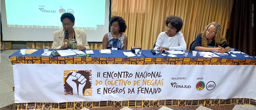 Imagem Acessível: A imagem mostra uma mesa de debates com quatro mulheres negras participando do II Encontro Nacional do Coletivo de Negras e Negros da Fenajud. Elas estão sentadas atrás de uma mesa coberta com tecido estampado e uma faixa que identifica o evento. Ao fundo, um telão projeta a identidade visual da atividade. O card traz o título: “SINJUS-MG reforça compromisso Antirracista em encontro nacional da FENAJUD”, destacando o compromisso da entidade com a pauta da igualdade racial.