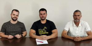 A imagem mostra três homens sentados lado a lado atrás de uma mesa, com uma bandeira do SINJUS MG estendida à frente. O homem ao centro, Fellipe Tomanini, novo diretor do SINJUS-MG, veste uma camiseta preta com a frase “Só a luta te garante” em amarelo, e segura uma caneta com papéis que oficializam o cargo. À esquerda e à direita, os dirigentes do sindicato Felipe Rodrigues e Alex Aguiar acompanham a cena. O card traz o título: “SINJUS institui Diretoria de Acessibilidade e Inclusão e empossa novo diretor”, reforçando a pauta de luta fortalecida do sindicato.