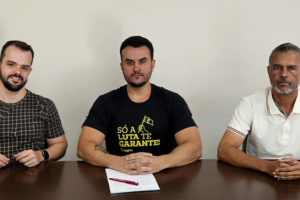 A imagem mostra três homens sentados lado a lado atrás de uma mesa, com uma bandeira do SINJUS MG estendida à frente. O homem ao centro, Fellipe Tomanini, novo diretor do SINJUS-MG, veste uma camiseta preta com a frase “Só a luta te garante” em amarelo, e segura uma caneta com papéis que oficializam o cargo. À esquerda e à direita, os dirigentes do sindicato Felipe Rodrigues e Alex Aguiar acompanham a cena. O card traz o título: “SINJUS institui Diretoria de Acessibilidade e Inclusão e empossa novo diretor”, reforçando a pauta de luta fortalecida do sindicato.