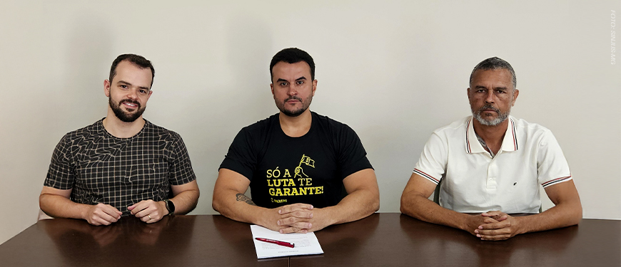 A imagem mostra três homens sentados lado a lado atrás de uma mesa, com uma bandeira do SINJUS MG estendida à frente. O homem ao centro, Fellipe Tomanini, novo diretor do SINJUS-MG, veste uma camiseta preta com a frase “Só a luta te garante” em amarelo, e segura uma caneta com papéis que oficializam o cargo. À esquerda e à direita, os dirigentes do sindicato Felipe Rodrigues e Alex Aguiar acompanham a cena. O card traz o título: “SINJUS institui Diretoria de Acessibilidade e Inclusão e empossa novo diretor”, reforçando a pauta de luta fortalecida do sindicato.