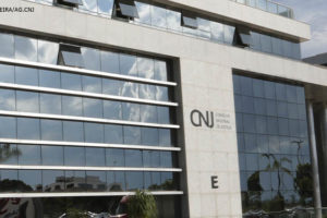 Fachada do edifício-sede do Conselho Nacional de Justiça (CNJ), em Brasília. Prédio retangular envidraçado com uma coluna central em concreto com o emblema do CNJ.