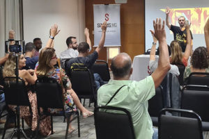 Auditório com pessoas em uma reunião (AGE), elas levantam as mãos em sinal de aprovação a uma proposta, à frente da plateia está Alexandre Pires, coordenador-geral do SINJUS-MG.