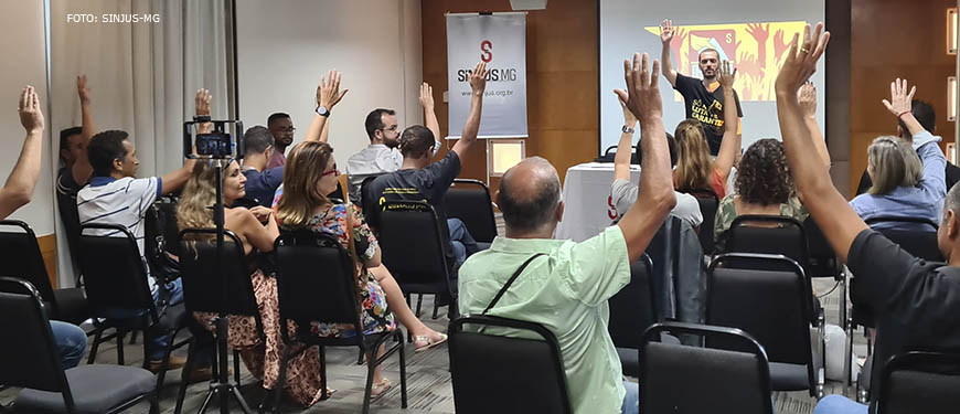Auditório com pessoas em uma reunião (AGE), elas levantam as mãos em sinal de aprovação a uma proposta, à frente da plateia está Alexandre Pires, coordenador-geral do SINJUS-MG.