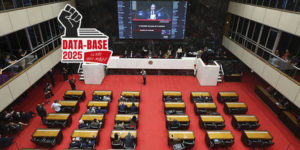 A imagem mostra o plenário da Assembleia Legislativa de Minas Gerais (ALMG) durante uma sessão, com deputados sentados e o telão exibindo a 1ª Reunião Solene do Plenário. O ambiente é imponente, com o tapete vermelho e o painel eletrônico em destaque. No canto superior direito, aparece o selo da campanha “Data-Base 2025 – Eu não abro mão!”, símbolo da mobilização sindical. Conteúdo textual: Pauta prioritária - SINJUS intensifica esforços pela Data-Base 2025 após início da sessão legislativa na ALMG.