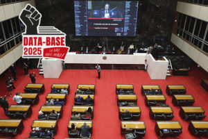 A imagem mostra o plenário da Assembleia Legislativa de Minas Gerais (ALMG) durante uma sessão, com deputados sentados e o telão exibindo a 1ª Reunião Solene do Plenário. O ambiente é imponente, com o tapete vermelho e o painel eletrônico em destaque. No canto superior direito, aparece o selo da campanha “Data-Base 2025 – Eu não abro mão!”, símbolo da mobilização sindical. Conteúdo textual: Pauta prioritária - SINJUS intensifica esforços pela Data-Base 2025 após início da sessão legislativa na ALMG.
