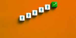 Imagem Acessível: A imagem traz uma composição com fundo alaranjado e uma fileira ascendente de blocos brancos com ícones de pessoas, finalizando com um bloco verde que exibe uma seta apontando para cima, simbolizando crescimento profissional e progressão na carreira. Conteúdo textual: Carreira - TJMG convoca candidatos à PV 2024 para retificação de documentos.