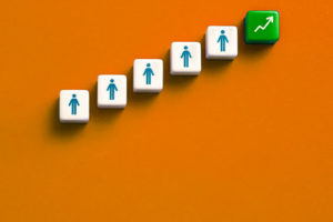 Imagem Acessível: A imagem traz uma composição com fundo alaranjado e uma fileira ascendente de blocos brancos com ícones de pessoas, finalizando com um bloco verde que exibe uma seta apontando para cima, simbolizando crescimento profissional e progressão na carreira. Conteúdo textual: Carreira - TJMG convoca candidatos à PV 2024 para retificação de documentos.