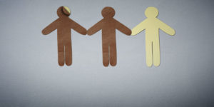 Imagem Acessível: A imagem traz três figuras humanas de papel, em cores diferentes, de mãos dadas, simbolizando diversidade racial. A primeira figura humana é de papel marrom, por baixo dela está uma figura em papel branco, a segunda figura é marrom e a terceira é bege claro. A imagem alerta para possíveis fraudes nas regras de fiscalização das candidaturas vinculadas às cotas raciais. Conteúdo textual: Eleições 2026 - Minuta do TSE ignora verificação racial obrigatória e coloca em risco política de cotas eleitorais.