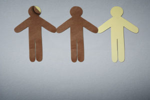 Imagem Acessível: A imagem traz três figuras humanas de papel, em cores diferentes, de mãos dadas, simbolizando diversidade racial. A primeira figura humana é de papel marrom, por baixo dela está uma figura em papel branco, a segunda figura é marrom e a terceira é bege claro. A imagem alerta para possíveis fraudes nas regras de fiscalização das candidaturas vinculadas às cotas raciais. Conteúdo textual: Eleições 2026 - Minuta do TSE ignora verificação racial obrigatória e coloca em risco política de cotas eleitorais.