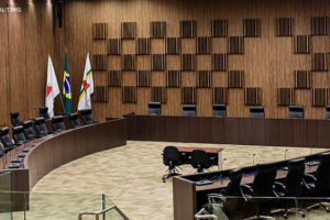 A imagem mostra o auditório onde são realizadas as sessões do Órgão Especial do TJMG. Há uma longa mesa disposta de forma quase circular com cadeiras preto. No fundo, há um painel de madeira e na lateral bandeiras do Brasil e Minas Gerais.
