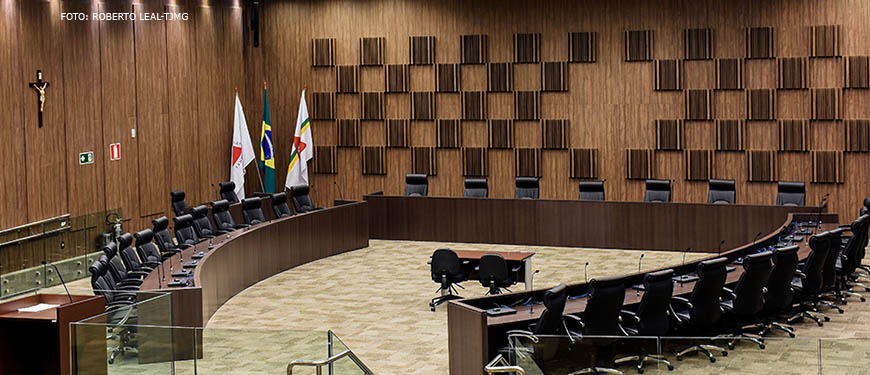 A imagem mostra o auditório onde são realizadas as sessões do Órgão Especial do TJMG. Há uma longa mesa disposta de forma quase circular com cadeiras preto. No fundo, há um painel de madeira e na lateral bandeiras do Brasil e Minas Gerais.