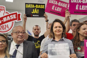 A imagem mostra um grupo de servidores e sindicalistas reunidos em um ambiente interno, segurando cartazes coloridos com mensagens de apoio à Data-Base 2024. Os cartazes exibem frases como “DATA-BASE JÁ!”, “DATA-BASE É LEI!” e “VOTE SIM – PL 3249/2025”. As expressões são de atenção, firmeza e esperança. No topo da imagem, há o selo “Data-Base 2024” com um punho erguido.