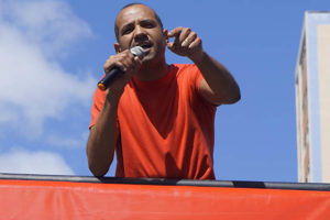 Homem negro de cabelos grisalhos (Wagner Ferreira) está sobre carro de som discursando com um microfone em sua mão.