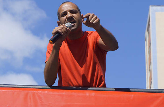 Homem negro de cabelos grisalhos (Wagner Ferreira) está sobre carro de som discursando com um microfone em sua mão.