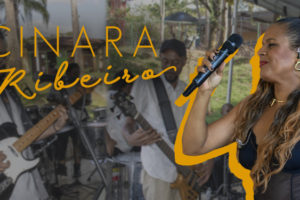 A imagem mostra a cantora Cinara Ribeiro em destaque, com microfone na mão e expressão vibrante durante uma apresentação. Ao fundo, sua banda toca ao ar livre em um ambiente descontraído. O texto informa que filiados ao SINJUS-MG têm 10% de desconto na contratação de shows da artista, por meio de um convênio exclusivo.