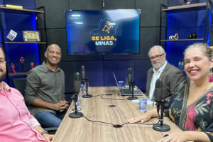 Imagem acessível: A imagem mostra o registo do episódio do podcast Se Liga, Minas, com quatro pessoas em um estúdio de gravação, sentadas ao redor de uma mesa com microfones. Ao fundo, uma TV exibe o logotipo do podcast. O texto em destaque informa: “Dirigentes do SINJUS debatem democracia e defesa dos servidores no Se Liga, Minas”.