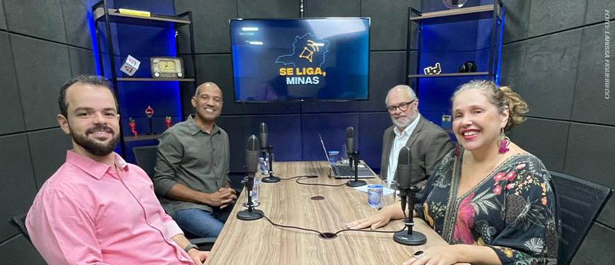 Imagem acessível: A imagem mostra o registo do episódio do podcast Se Liga, Minas, com quatro pessoas em um estúdio de gravação, sentadas ao redor de uma mesa com microfones. Ao fundo, uma TV exibe o logotipo do podcast. O texto em destaque informa: “Dirigentes do SINJUS debatem democracia e defesa dos servidores no Se Liga, Minas”.