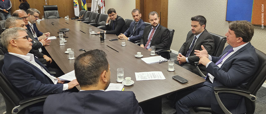 No card há dois registros fotográficos da Mesa de Negociações, as imagens mostram ângulos opostos de uma mesa de reunião em madeira escura, ao redor dela estão homens com trajes formais sentados. Sobre a mesa, copos d'água, xícaras de café e documentos. Ao fundo, uma televisão desligada e um quadro abstrato nas cores azul, amarelo e laranja. nesta reunião estão representantes da presidência do TJ, dirigentes sindicais do Serjusmig, Sindojus/MG, representando o SINJUS-MG estão o coordenador-geral, Felipe Rodrigues, o diretor administrativo, Alexandre Pires e o diretor de Finanças, Alex Aguiar.
