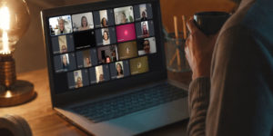 Imagem Acessível: A imagem mostra uma pessoa sentada diante de um notebook em um ambiente interno, iluminado por luz quente. Ela segura uma caneca escura enquanto acompanha uma videoconferência. Na tela do computador aparecem diversas participantes em uma reunião virtual, mediada pela diretora Patrícia Oliveira, as mulheres estão organizadas em uma grade com câmeras ligadas e desligadas. Conteúdo textual: MULHERES QUE CORREM COM OS LOBOS - Círculo do Livro promove reflexão sobre relacionamentos e dualidade feminina.