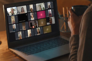Imagem Acessível: A imagem mostra uma pessoa sentada diante de um notebook em um ambiente interno, iluminado por luz quente. Ela segura uma caneca escura enquanto acompanha uma videoconferência. Na tela do computador aparecem diversas participantes em uma reunião virtual, mediada pela diretora Patrícia Oliveira, as mulheres estão organizadas em uma grade com câmeras ligadas e desligadas. Conteúdo textual: MULHERES QUE CORREM COM OS LOBOS - Círculo do Livro promove reflexão sobre relacionamentos e dualidade feminina.