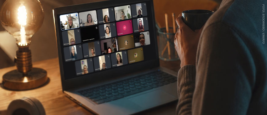 Imagem Acessível: A imagem mostra uma pessoa sentada diante de um notebook em um ambiente interno, iluminado por luz quente. Ela segura uma caneca escura enquanto acompanha uma videoconferência. Na tela do computador aparecem diversas participantes em uma reunião virtual, mediada pela diretora Patrícia Oliveira, as mulheres estão organizadas em uma grade com câmeras ligadas e desligadas. Conteúdo textual: MULHERES QUE CORREM COM OS LOBOS - Círculo do Livro promove reflexão sobre relacionamentos e dualidade feminina.