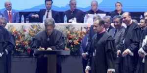 A imagem mostra uma cerimônia oficial no Grande Teatro do Palácio das Artes em Belo Horizonte.. Em primeiro plano, um desembargador veste toga preta e assina um documento em um púlpito (o novo presidente do TJMG, Vicente de Oliveira Silva, enquanto outro magistrado (o desembargador José Carlos Corrêa Junior), também de toga, observa ao lado. Ao fundo, há uma mesa de autoridades, com magistrados e representantes institucionais sentados, além de outras pessoas em pé acompanhando a solenidade. O palco é decorado com arranjos de flores em tons de laranja, vermelho e amarelo.