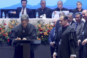 A imagem mostra uma cerimônia oficial no Grande Teatro do Palácio das Artes em Belo Horizonte.. Em primeiro plano, um desembargador veste toga preta e assina um documento em um púlpito (o novo presidente do TJMG, Vicente de Oliveira Silva, enquanto outro magistrado (o desembargador José Carlos Corrêa Junior), também de toga, observa ao lado. Ao fundo, há uma mesa de autoridades, com magistrados e representantes institucionais sentados, além de outras pessoas em pé acompanhando a solenidade. O palco é decorado com arranjos de flores em tons de laranja, vermelho e amarelo.