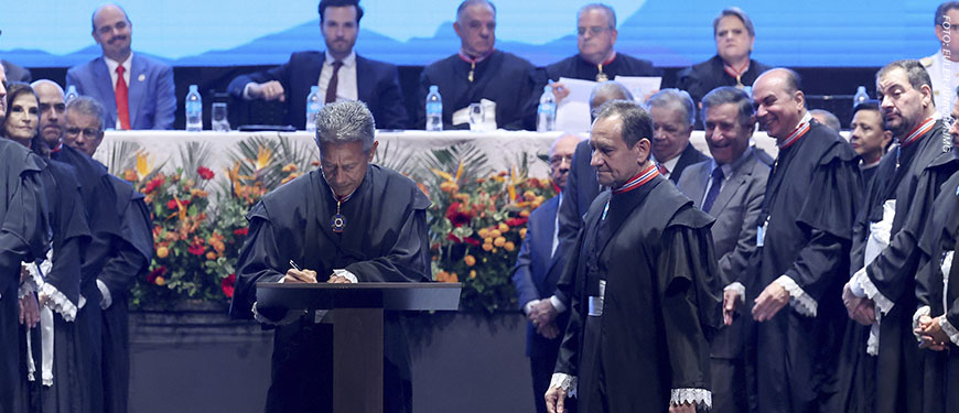 A imagem mostra uma cerimônia oficial no Grande Teatro do Palácio das Artes em Belo Horizonte.. Em primeiro plano, um desembargador veste toga preta e assina um documento em um púlpito (o novo presidente do TJMG, Vicente de Oliveira Silva, enquanto outro magistrado (o desembargador José Carlos Corrêa Junior), também de toga, observa ao lado. Ao fundo, há uma mesa de autoridades, com magistrados e representantes institucionais sentados, além de outras pessoas em pé acompanhando a solenidade. O palco é decorado com arranjos de flores em tons de laranja, vermelho e amarelo.