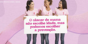 Imagem Acessível: Grupo de três mulheres (uma branca, uma negra e outra asiática), todas vestem calça jeans, com camiseta rosa claro e usam a fita cor de rosa que simboliza o combate ao câncer de mama. Elas carregam um cartaz com a mensagem: O câncer de mama não escolhe idade, mas podemos escolher a prevenção.