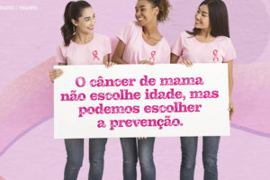 Imagem Acessível: Grupo de três mulheres (uma branca, uma negra e outra asiática), todas vestem calça jeans, com camiseta rosa claro e usam a fita cor de rosa que simboliza o combate ao câncer de mama. Elas carregam um cartaz com a mensagem: O câncer de mama não escolhe idade, mas podemos escolher a prevenção.