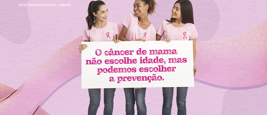 Imagem Acessível: Grupo de três mulheres (uma branca, uma negra e outra asiática), todas vestem calça jeans, com camiseta rosa claro e usam a fita cor de rosa que simboliza o combate ao câncer de mama. Elas carregam um cartaz com a mensagem: O câncer de mama não escolhe idade, mas podemos escolher a prevenção.