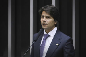 A imagem mostra o Deputado Pedro Paulo de terno falando ao microfone em um ambiente formal.